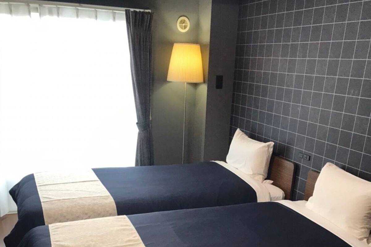 A hotel room with two beds, a nightstand, a lamp, and a view of the outdoors.