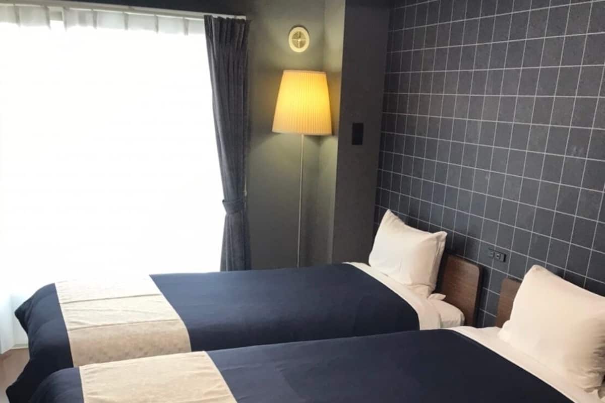A hotel room with two beds, a nightstand, a lamp, and a view of the outdoors.