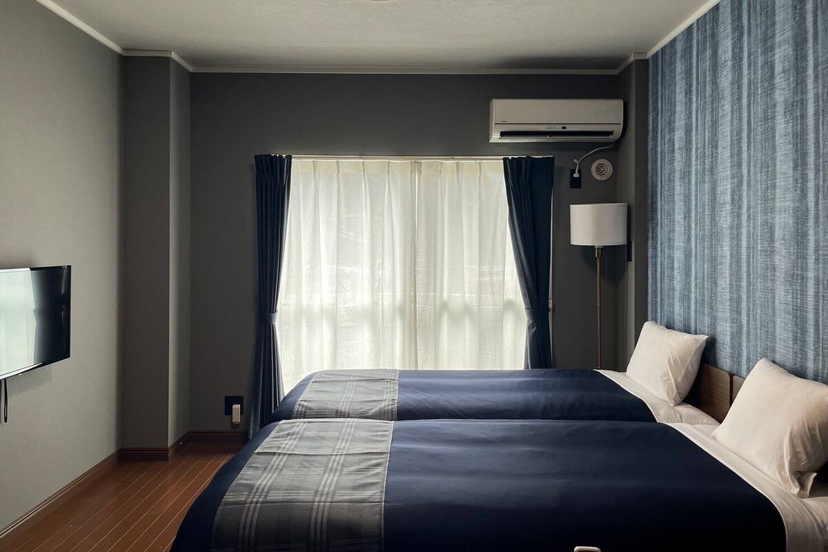 A hotel room with a bed, a flat-screen TV, an air conditioner, and a window with curtains.