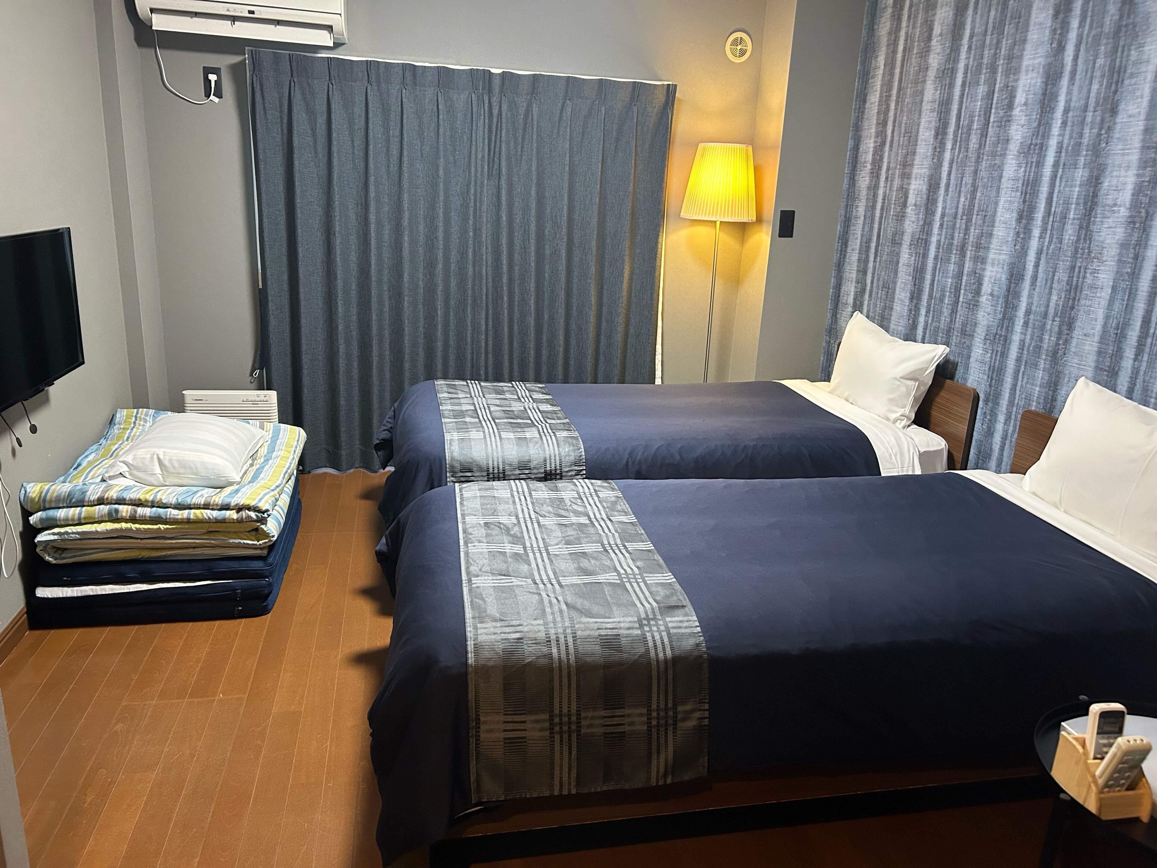 A hotel room with two beds, a flat-screen TV, an air conditioner, a lamp, and a bedside table with a phone.