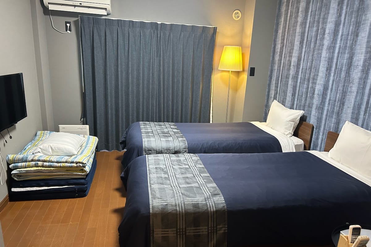 A hotel room with two beds, a flat-screen TV, an air conditioner, a lamp, and a bedside table with a phone.