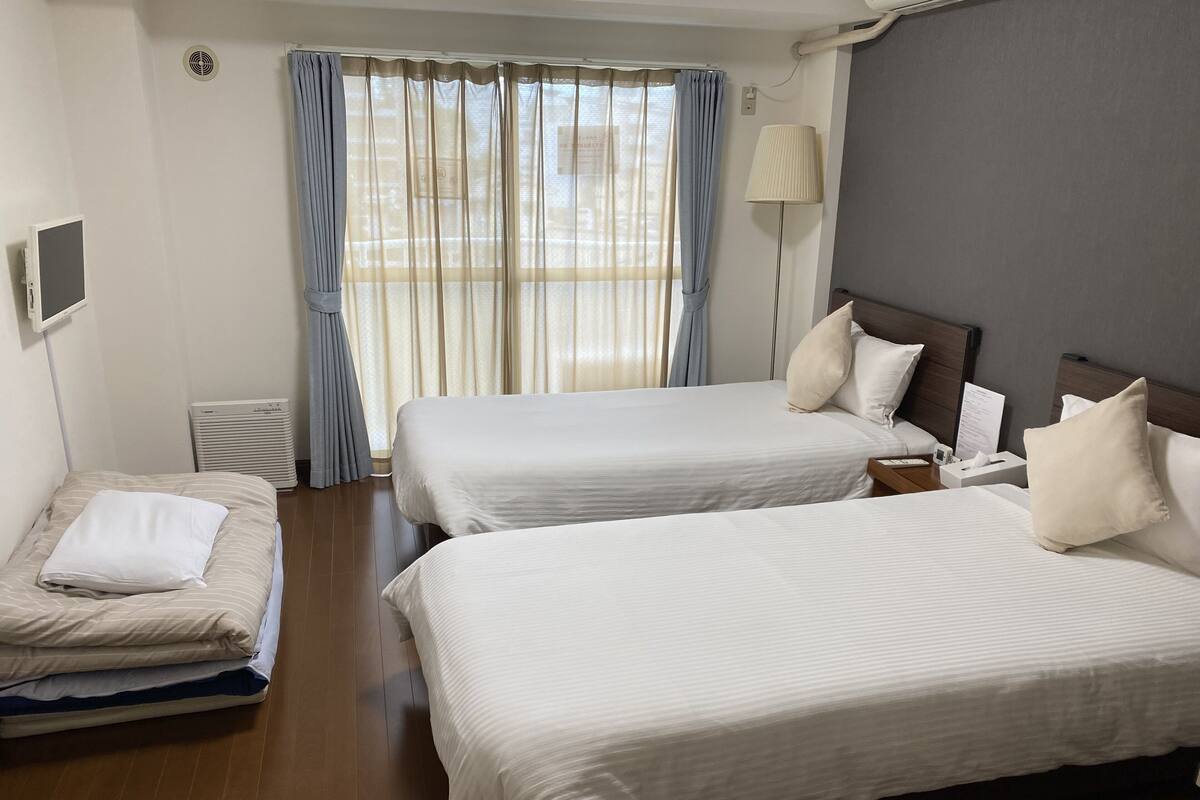 A hotel room with two beds, a bedside table, a lamp, and a window with curtains.