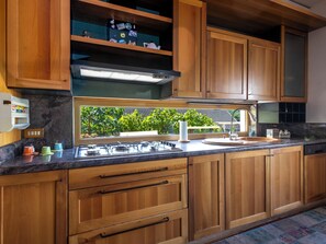 Cocina con armarios de madera, estufa a gas, lavamanos y ventana con vista a vegetación.