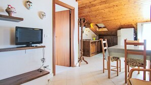 Ein gemütliches Zimmer mit Holzbalkendecke, einem Fernseher auf einem Regal, einem Esstisch mit Stühlen und einer Küchenzeile mit Kühlschrank.