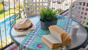 Ein runder Tisch mit einem Blumentopf, einem offenen Buch, einem Hut und einer Tasse auf einer gemusterten Tischdecke.