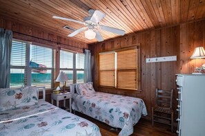 Una cabaña con dos camas, un ventilador de techo, una ventana con persianas y una lámpara en una mesita de noche.