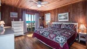 Une chambre avec un plafond en bois, un lit recouvert d’une courtepointe à motifs, une commode et une vue sur l’océan par une porte vitrée.
