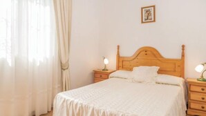 Een net opgemaakt bed met een houten hoofdeinde, twee nachtkastjes met lampen, en een enkel fotolijstje aan de muur.
