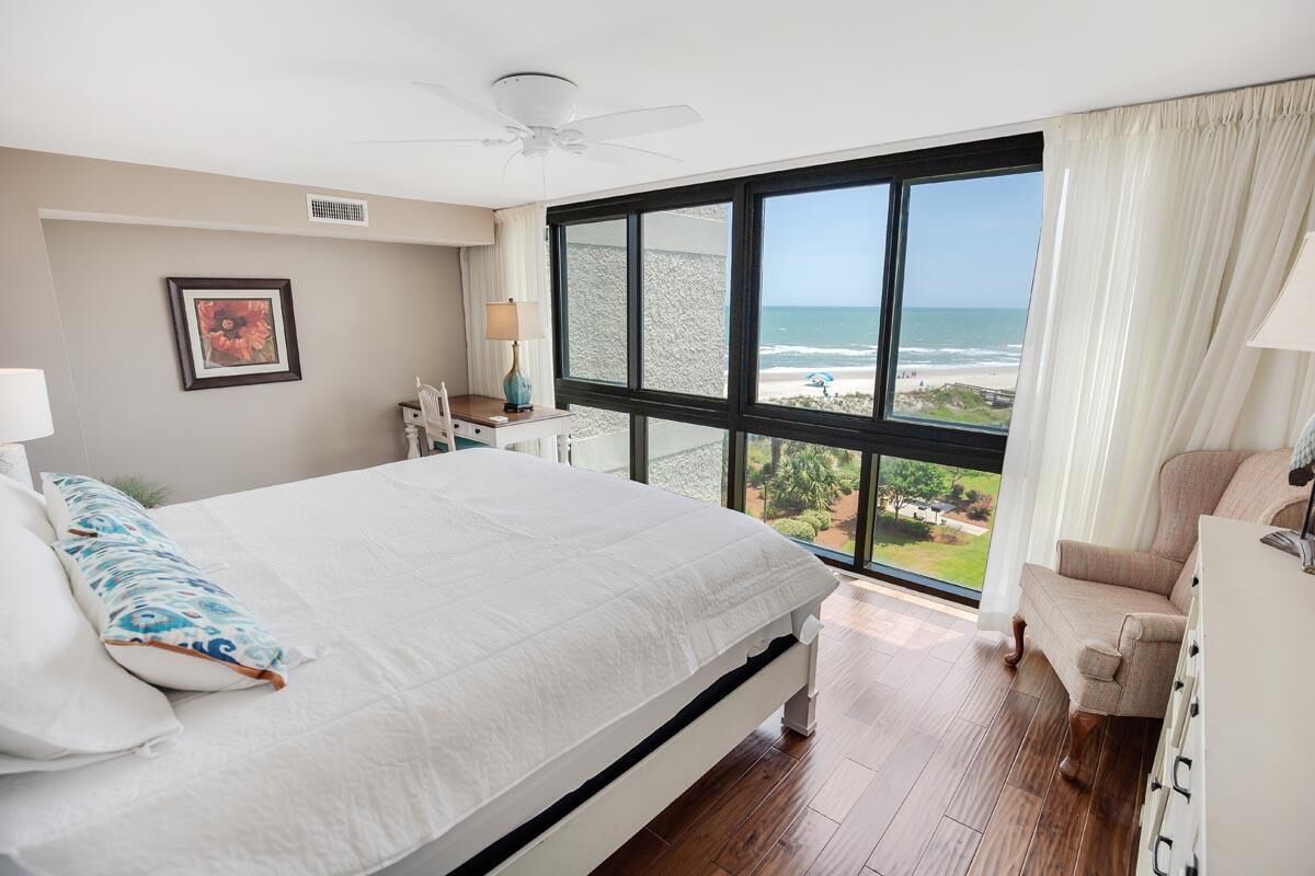 A bedroom with a large bed, a chair, a desk, and a view of the beach.