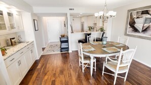 A dining area with a wooden table and white chairs, a chandelier, and a kitchen in the background. - Paradise Found Beachfront Litchfield By the Sea (Pawleys Island)