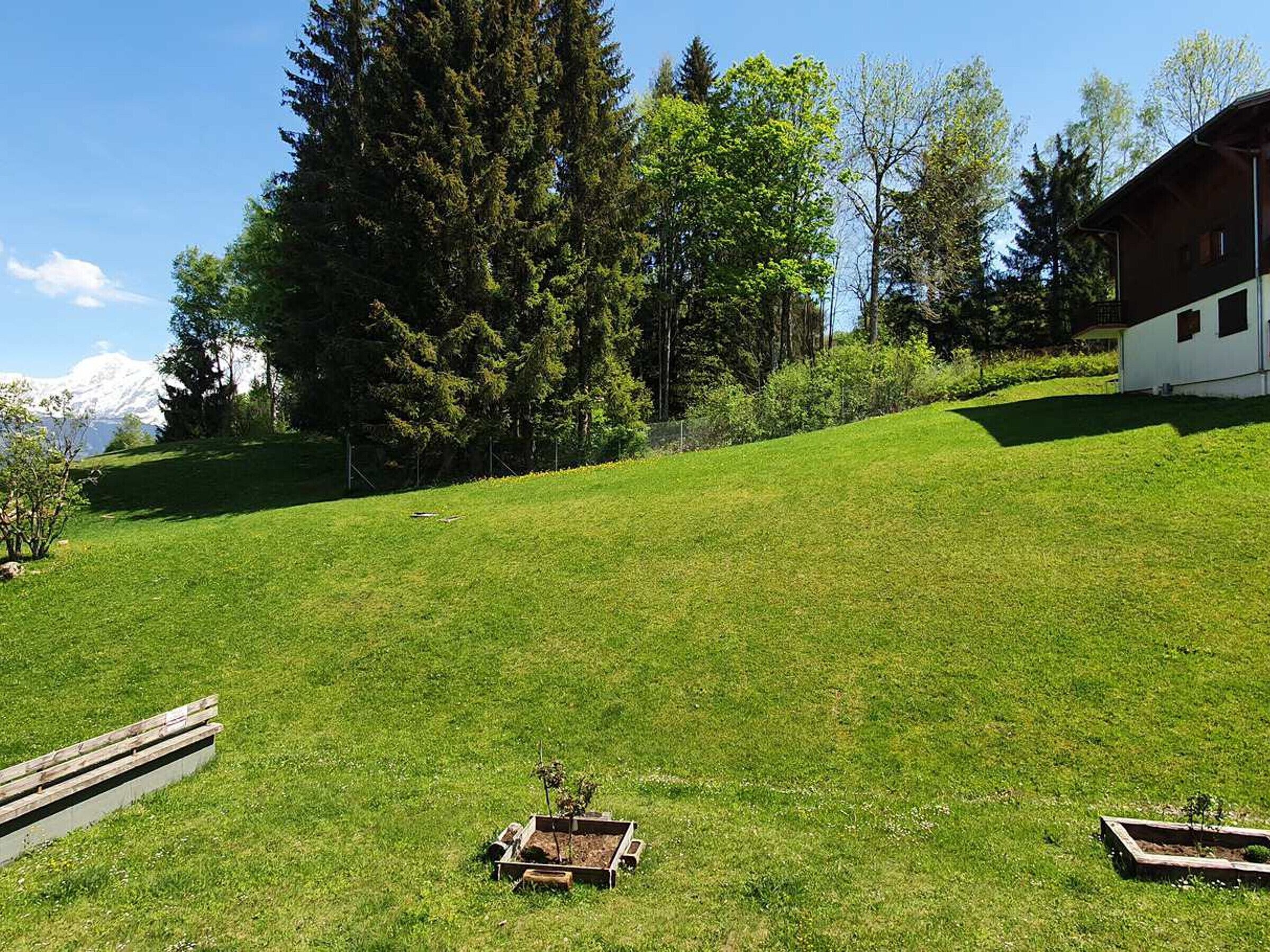 Un campo cubierto de césped con un banco de madera y jardines elevados, una cabaña de madera, y un fondo de árboles y una montaña distante.