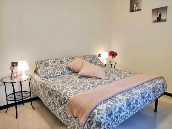 A bedroom with a floral-patterned bedspread, a single pink pillow, a knitted throw blanket, and two bedside tables with lamps and decorative items.