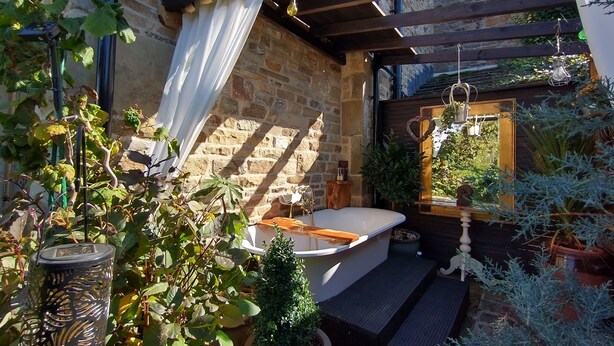 Une salle de bain extérieure rustique, dotée d’une baignoire sur pieds, entourée de plantes en pot et d’une pergola en bois.
