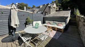 En træterrasse med et rundt bord og stole, et blødt siddeområde og en lille overdækket plads med skrå tag.