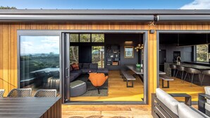 A modern interior with a wooden exterior, a dining area with a bench, a sofa, and a kitchen with bar stools. - Brand new luxury lodge in Onetangi, with pool, spa and sleeping 10 (Waiheke Island)