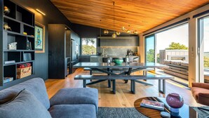 A modern living room with a wooden ceiling, a dining area with a large table, and a kitchen with a view. - Brand new luxury lodge in Onetangi, with pool, spa and sleeping 10 (Waiheke Island)