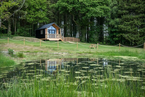 Eine Hütte mit einer Terrasse, umgeben von einem Seilzaun, in der Nähe eines Teichs mit Seerosen.