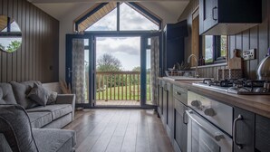 A modern kitchen with a large window, a built-in oven, and a dining area with a wooden table and chairs. - Cabin In The Woods - 1 Bed - Kilgetty (Kilgetty)