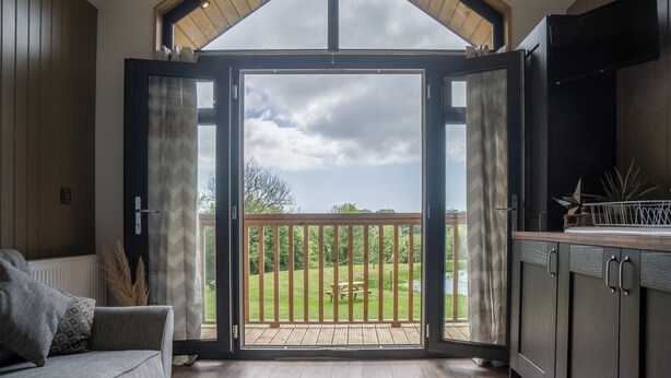 A room with a large sliding glass door leading to a balcony with a wooden deck and a picnic table. - Cabin In The Woods - 1 Bed - Kilgetty (Kilgetty)