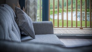 A grey sofa with patterned cushions in a room with a large window overlooking a wooden deck and a body of water. - Cabin In The Woods - 1 Bed - Kilgetty (Kilgetty)