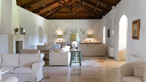 A spacious room with a high ceiling, white walls, and a tiled floor. There are several white sofas and a small table with a lamp.