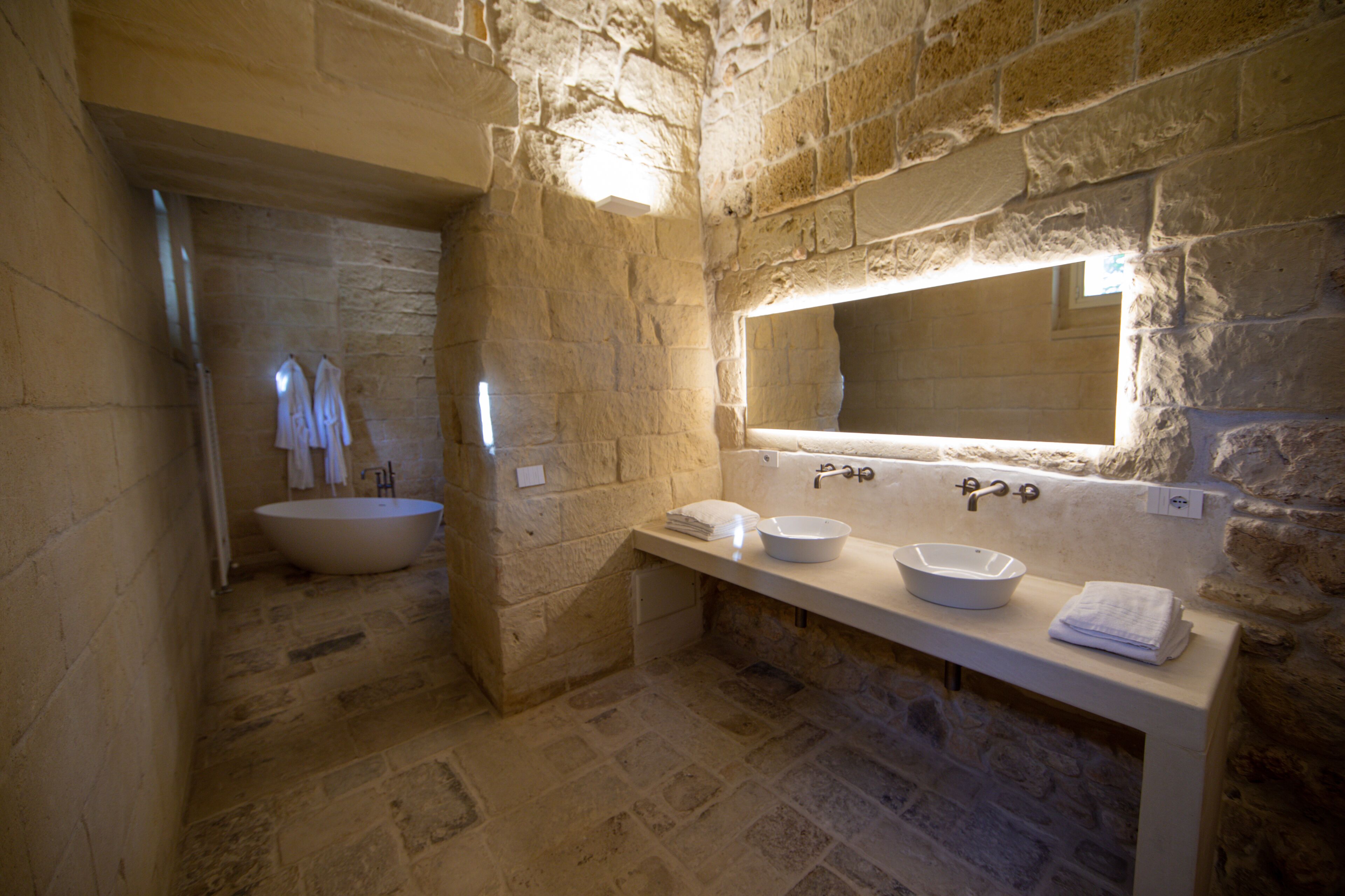 A stone-walled bathroom with a freestanding bathtub, a double sink with a bowl, and a mirror above the sink.