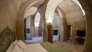 A room with a bed, a television, and a chair. The room has stone walls and arches.