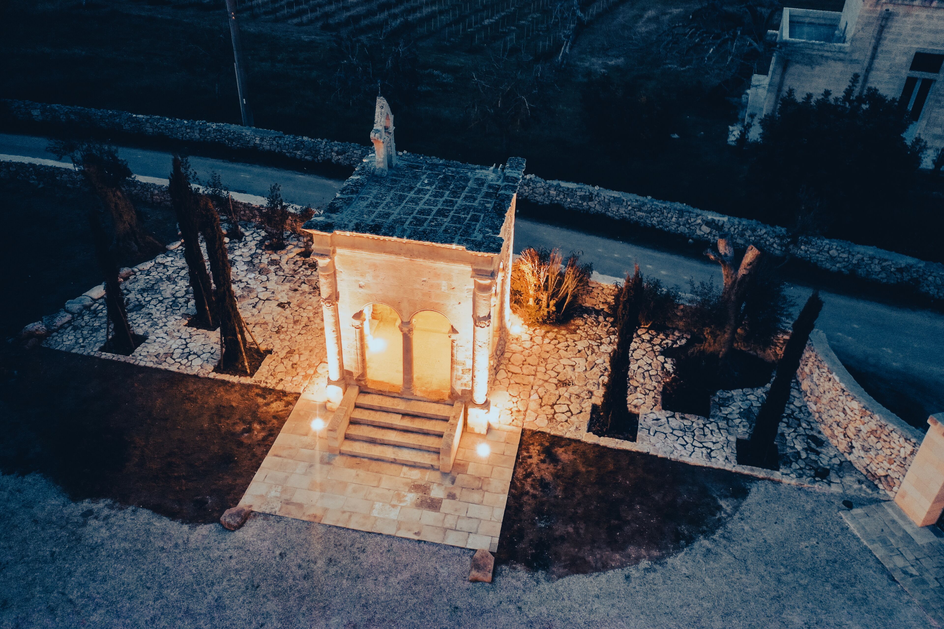 Una pequeña estructura iluminada con techo de tejas y escalones que llevan a ella, rodeada de árboles y un muro de piedra.