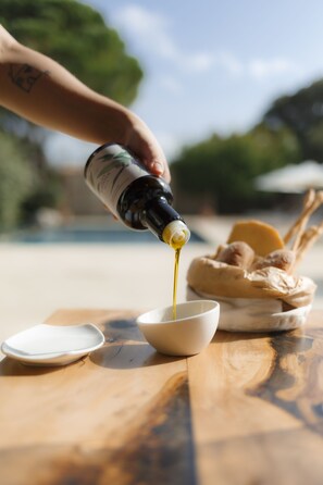 A hand pouring olive oil into a bowl on a wooden table with bread and a plate. - Masseria Lilei - Puglia, Salento (Lizzanello)