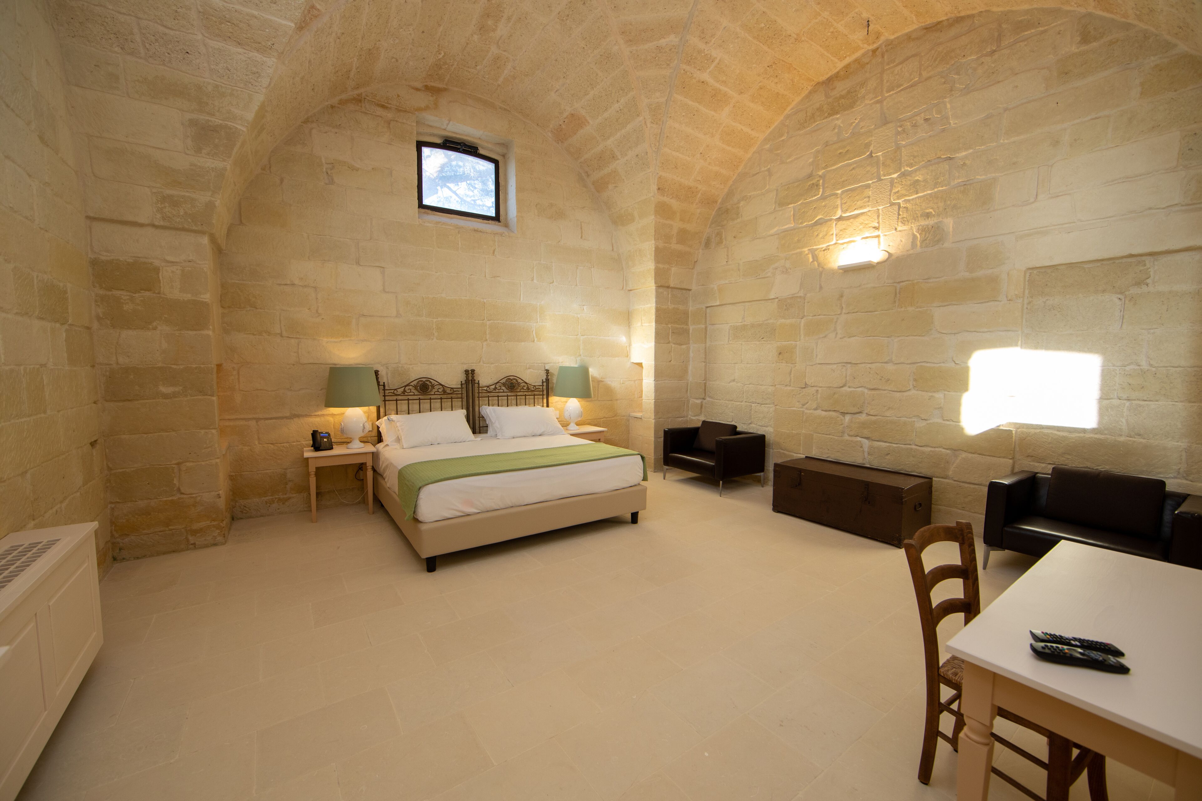 A stone-walled room with a bed, bedside lamps, a small table, and a chair.