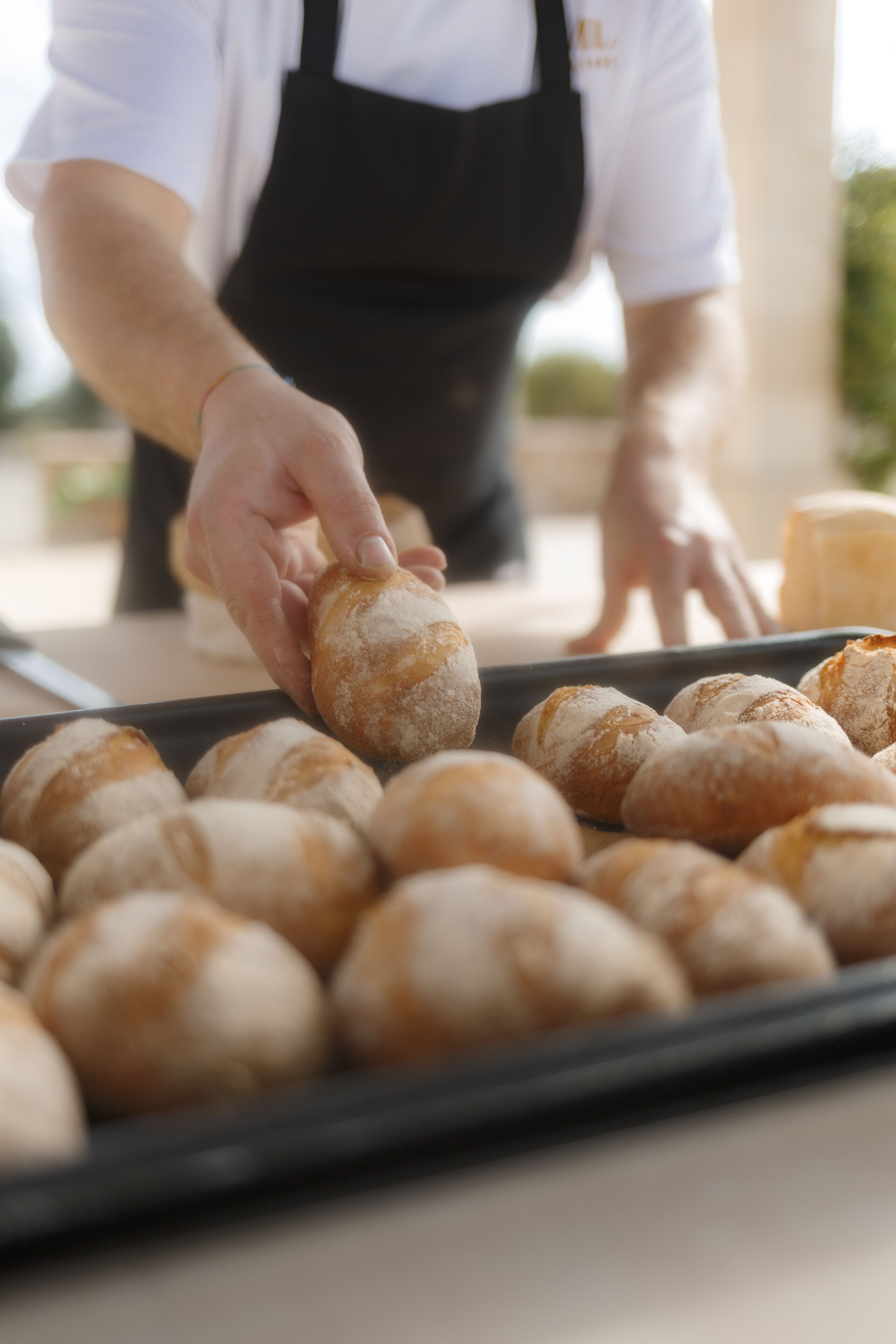 A person in a white shirt and black apron handling bread rolls on a tray.