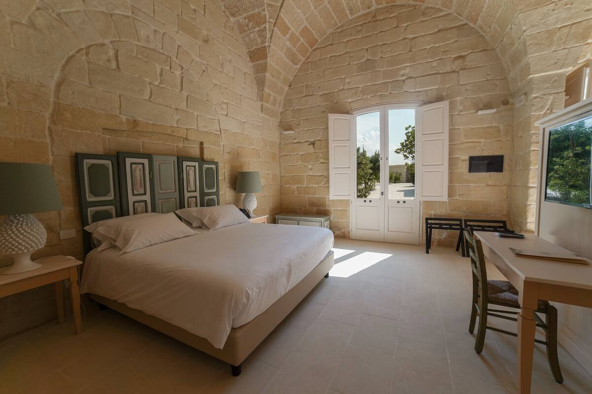Ein Schlafzimmer mit einem großen Bett, einem Schreibtisch und einem Stuhl. Ein Fenster bietet Blick auf Bäume.