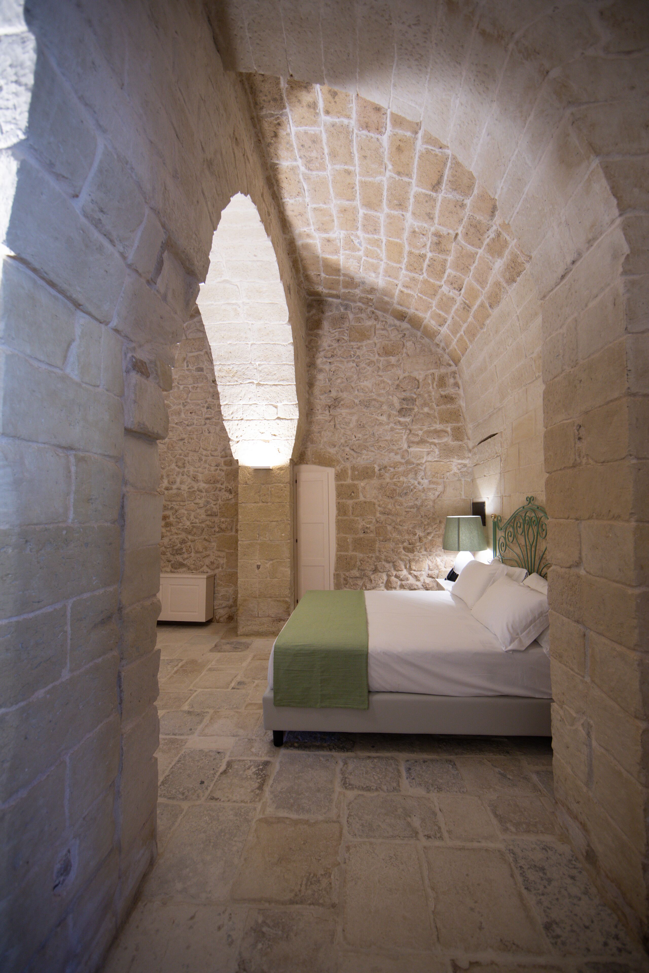 A bedroom with a stone archway, a bed with a green blanket, a bedside table, and a lamp.