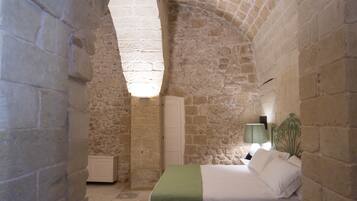 A bedroom with a stone archway, a bed with a green blanket, a bedside table, and a lamp.