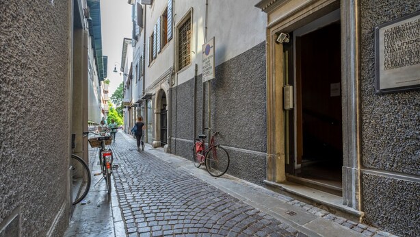 A cobblestone street with buildings on either side, a bicycle, and a person walking. - GuestHost - The property enjoys a strategic position, just a few steps from the historic center and only 10 minutes by car from the hospital. Its location makes the apartment perfect for any type of trip: nearby, you can find typical bars, ethnic restaur (Udine)