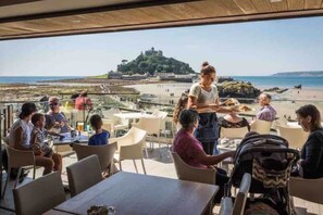 Un restaurant avec une terrasse offrant une vue sur une plage et un château.