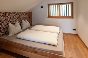Un dormitorio con cabecera de madera, cama con ropa de cama blanca, ventana con persianas verticales y una lámpara de pared pequeña.