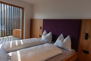 A double bed with white linens and pillows, a wooden headboard, and a view through a large window.