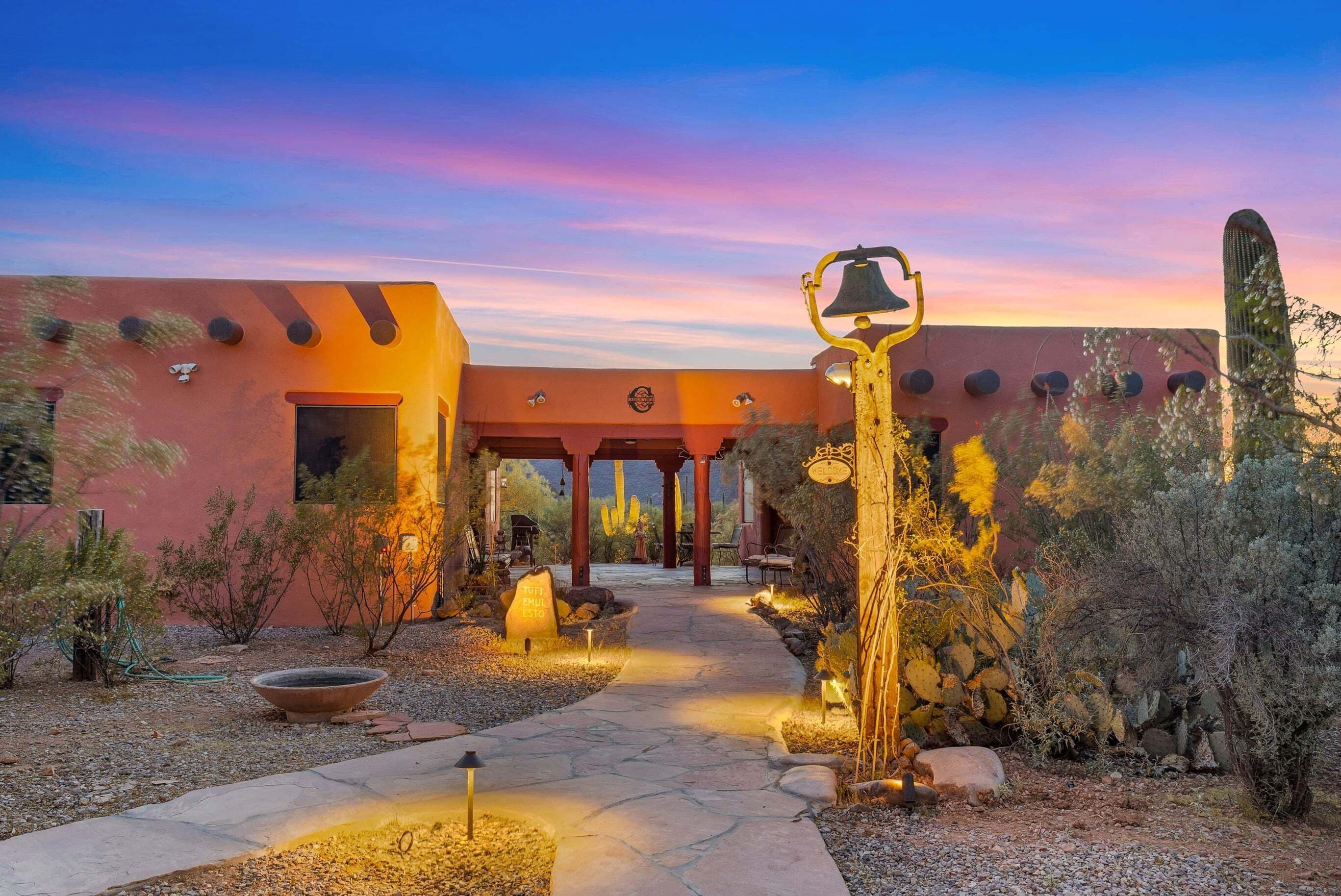 A desert-style building with a stone pathway leading to a covered entrance, surrounded by cacti and desert plants.