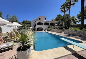 Zona de piscina con un edificio blanco al fondo, rodeada de palmeras y un muro de piedra.