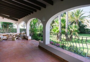 Un patio techado con una mesa y sillas, vista a un jardín cuidado con palmeras y una barandilla de metal.