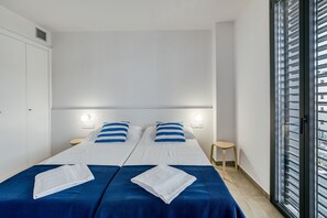 Ein Hotelzimmer mit zwei Betten, einem hölzernen Hocker, einem Fenster mit Jalousien und einem weißen Deckenventilator.