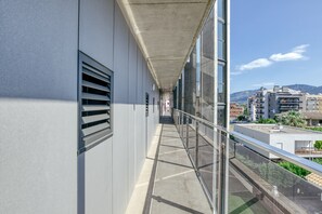 A corridor with a glass railing, leading to a balcony with a view of buildings and mountains.