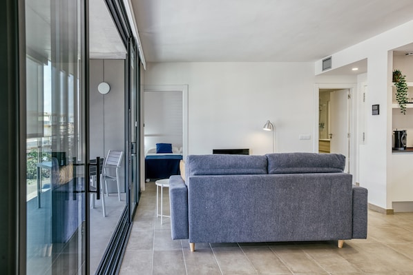 A modern living room with a grey sofa, a small table, and a chair. There is a glass wall with a view of the outside.