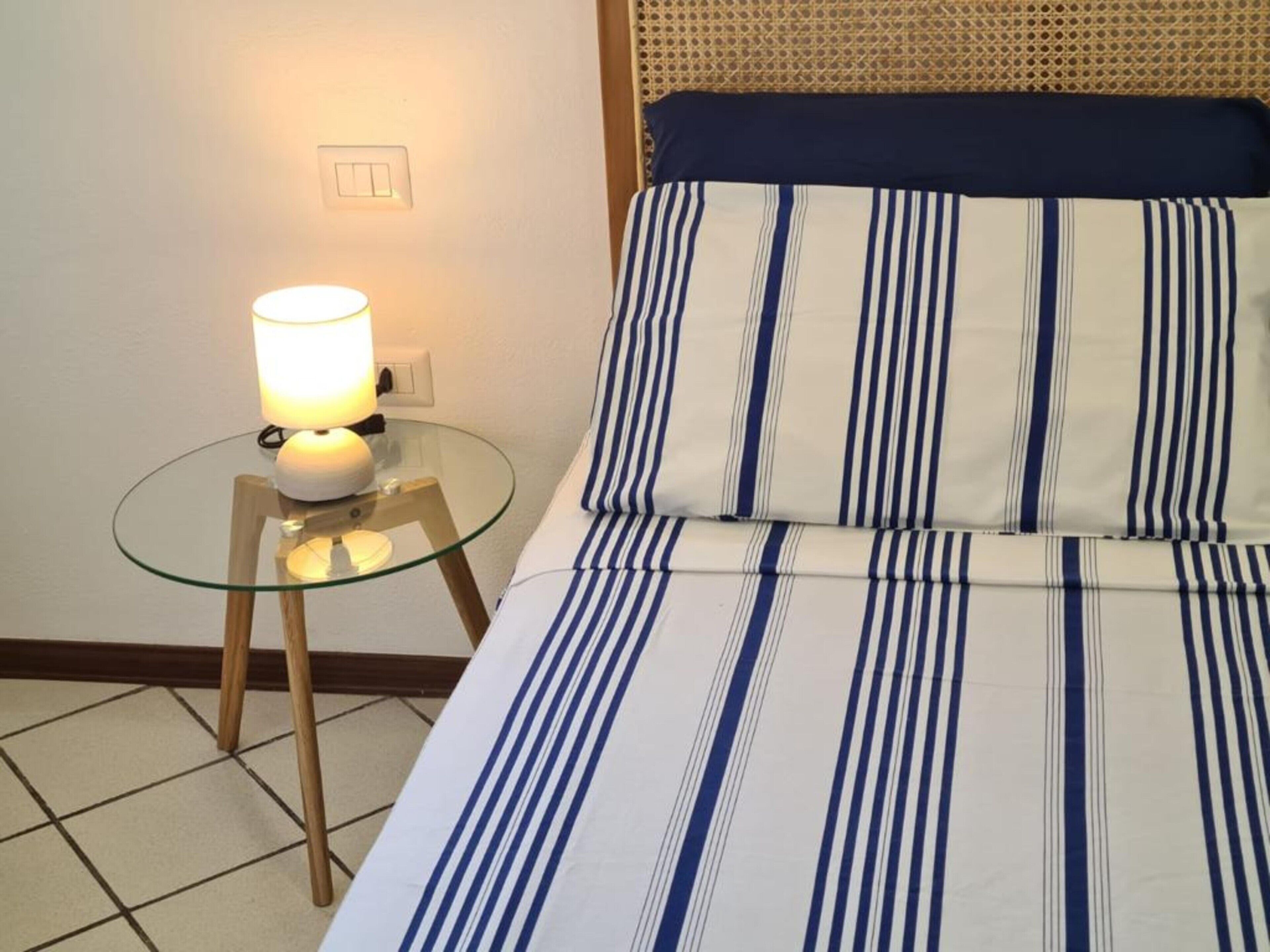 Una cama con colcha a rayas, una lámpara de noche sobre una mesita de cristal y un interruptor de pared.