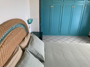 A bedroom with a wicker headboard, a bed with grey bedding, a small table, and a teal-colored wardrobe with louvered doors.