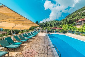 Una piscina con sillones de descanso y una zona techada, rodeada de vegetación y edificios.