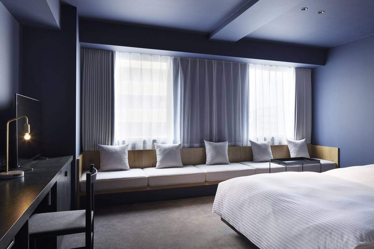 A modern hotel room with a large bed, a desk with a lamp, a bench with cushions, and large windows with curtains.