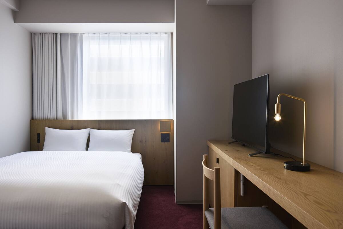 A hotel room with a bed, a desk with a TV, and a window with curtains.