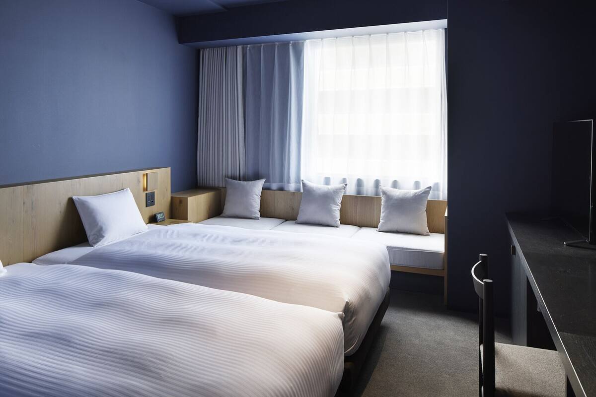 A hotel room with two beds, a large window with curtains, a bedside table, and a nightstand.